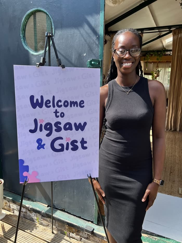 Bethany, smiling, next to the Welcome to Jigsaw & Gist sign